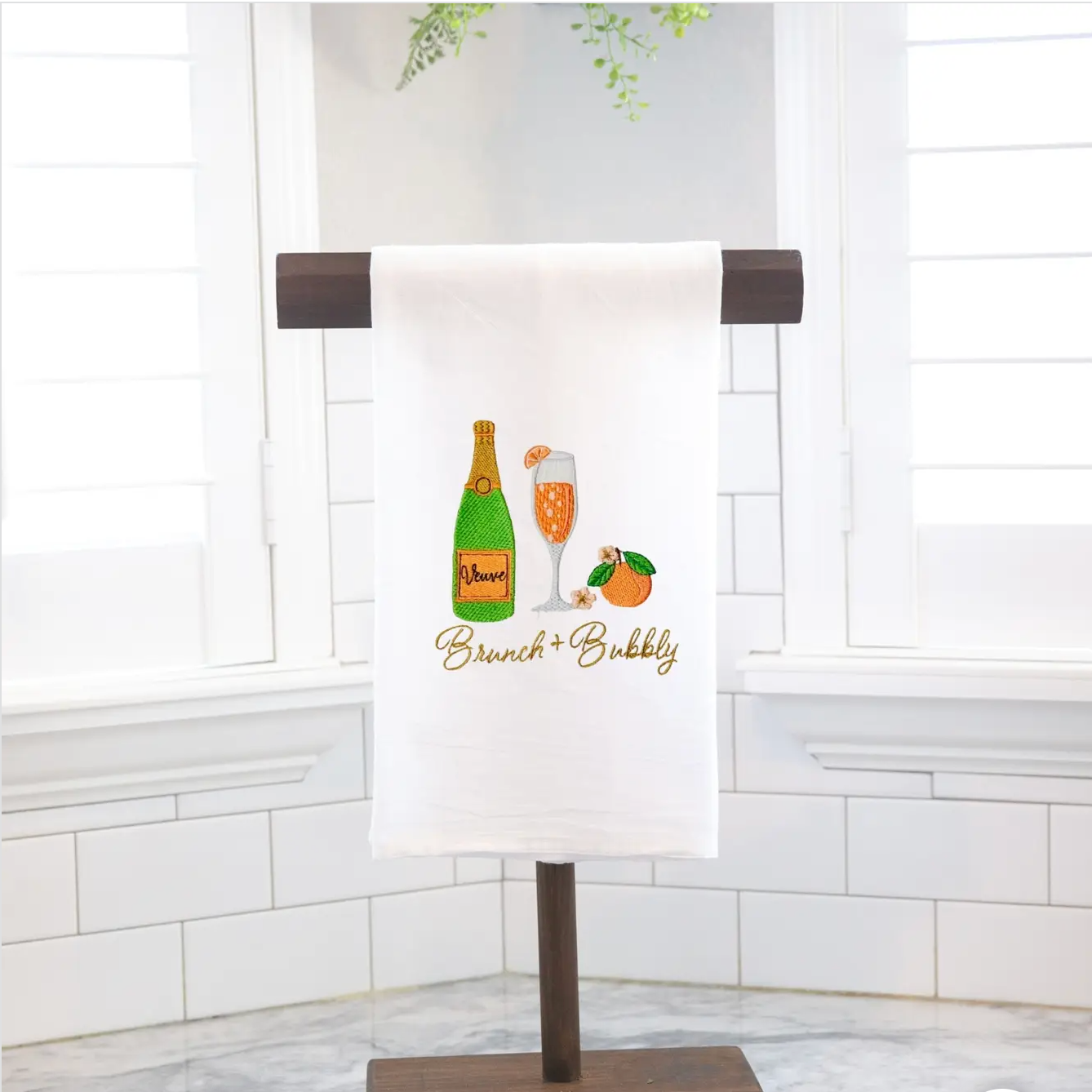 Tea Towel hanging on a wooden stand. The towel says "Brunch + Bubbly" and has an image of champagne, a glass, and an orange